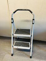 Front view of the 2 step foldable ladder showing steps and handle, placed on grey wood flooring against a beige wall.
