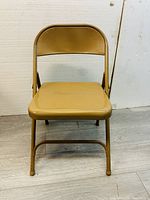 Front view of a tan metal foldable chair, showing padded seat and backrest with metal legs.