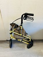Side view of a foldable mobility aid walker with yellow frame, ergonomic black handles with brakes, and a checkered fabric bag attached.