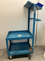 Full view of the blue metal order picker rolling cart showing two shelves and handle with visible wear and paper sticky notes attached.