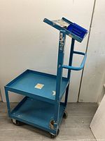 Side view of blue metal rolling cart showing two flat shelves, a tubular handle, and front wheels.