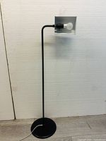Black floor lamp with minimalist design against a plain wall, showing the bulb socket and base with power cord on the floor.