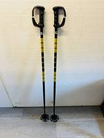 Full length front view of two ski poles standing on a floor against a light wall. The poles have black and yellow striped shafts and black handles with hand guards.
