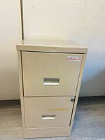 Front view of a beige metal storage/file cabinet with two drawers, showing wear and lock with no key.