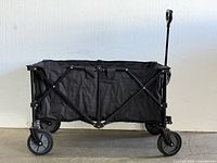 Side view of the black foldable utility cart showing its fabric body, metal frame with cross braces, and four wheels including two larger front wheels. The cart is unfolded and ready for use.
