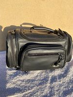 Black soft carrying case with zippered compartments shown from side with top handle visible.