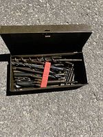 Open metal toolbox showing a variety of large and small metal twist drill bits inside, some bundled.