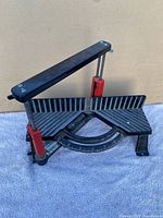 Full view of the miter saw angle guide showing black metal body and red clamps, resting on fabric background.