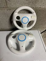 Photo showing two white Nintendo Wii steering wheels, one upright and one laying flat to display the Wii logo and button details.
