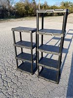 Two black plastic storage shelves, one with four tiers and one with three, shown standing outside on concrete surface with trees in the background.