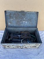 Open metal toolbox showing various clamps inside on a fabric surface.