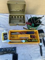 Overview of tools arranged on white cloth against a plain wall; includes tool trays with multiple small hand tools, a metal toolbox, oil bottle, and paint brushes.