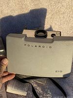 Photo showing the closed gray carrying case for the Polaroid Land Camera 210 with black text and lens detail on top.
