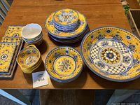 Full set of 26-piece Le Cadeaux dinnerware displayed on wooden table showing assortment of plates, bowls, and serving dishes in yellow and blue floral Mediterranean design.