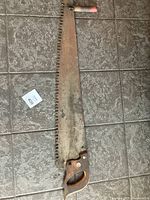 Full length view of the antique two-man crosscut saw lying flat on tiled floor. Shows steel blade with rust, large teeth, wooden handle with metal rivets and auxiliary top handle with red paint worn off.