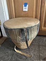 Full view of the medium vintage African drum showing natural animal skin drumhead, woven rope securing the head, and wooden body with broken leather strap lying on floor.