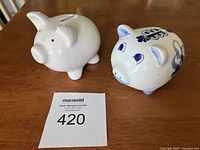 Photo of the two ceramic piggy banks on a wooden surface, showing the plain white one and the blue windmill patterned one.