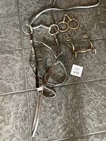 Photo showing brown leather horse bridle with reins alongside three metal horse bits on tiled floor.