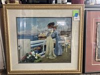 Framed artwork showing two women standing by the sea on a balcony with flowers and ships in the background. The frame is wooden with gold finish.