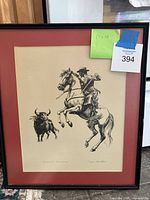 Full view of framed lithograph showing bullfighter on horseback against a bull, framed with red matting and black frame.