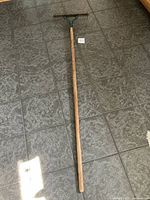 Full view of a long garden cultivator with wooden handle and metal blade head lying on a tiled floor.