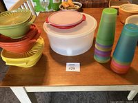Set of round Tupperware plastic containers with lids in various colors and two stacks of multicolor plastic cups on a wooden table.