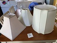 Three fabric lamp shades displayed on a wooden table: hexagonal, round, and square shapes in white and cream tones