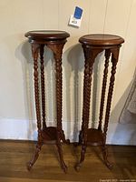 Pair of vintage wooden pedestal plant stands shown against a wall, dark brown in color, featuring spiral carved legs and claw feet with small lower shelves.