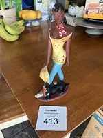 Front and side views of the ceramic figurine depicting a woman standing with a dog at her feet on a brown base. The woman is dressed in a yellow top, blue pants, white shoes, and a red and white scarf, holding a yellow fan.