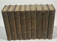 Front view of all nine antique brown leather volumes, standing upright, showing spine wear and embossed decoration.
