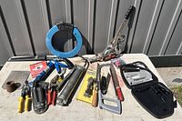 Wide view of all tools displayed on a table outside, showing hand winch, hacksaw, grease gun, clamps, wire cutters, pliers, utility knife, and blue strap among others.