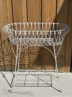 Full frontal image of antique white metal plant stand showing upper basket with wire loops and lower shelf with metal slats against a wooden fence background.