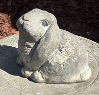 Gray resin bunny rabbit figurine shown outdoors on stone surface with mulch background, front and side view.