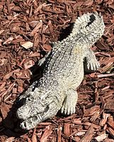Concrete alligator garden statue lying on bark mulch showing top detail and texture of scales and limbs, outdoor wear effects visible