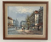 Framed oil on canvas painting showing a street scene in Paris with multiple buildings, pedestrians, and a domed building in the background. The painting is visible with wooden frame and fabric inner border.