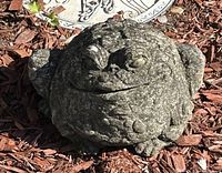 Front view of chubby concrete toad statue placed outdoors on mulch ground with a decorative plaque in the background.