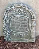 Concrete wall plaque photographed outdoors on mulch, showing detailed poem inscription and decorative floral and scroll elements.