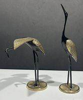 Pair of black enamel and brass crane bird statues on round brass bases against gray background.