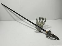 Full length side view of the metal sword showing blade and guard design on a hand-shaped stand.