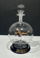 Front view of clear glass decanter with suspended Tiger Moth airplane in gold and purple glass and round stopper on top, mounted on black base with label.