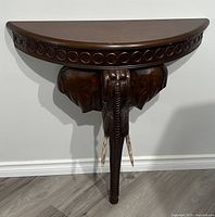Full view of half-moon shaped wood hall table supported by carved elephant head and trunk with carved pattern on top edge