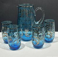 Front and side view of blue glass pitcher with clear twisted handle and gold floral decoration, with four matching blue glass tumblers having the same decorative pattern