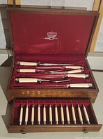 Complete open wooden box showing upper section with carving knives and tools with ivory-colored handles and lower drawer with twelve matching forks.