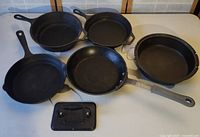 Top view of set of five cast iron pans and rectangular lid on white surface.