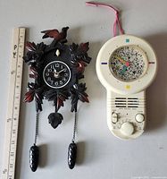 Pair of clocks displayed side by side with ruler for scale. Smaller dark wood-tone cuckoo clock with chains and decorative bird on left, white circular retro clock with multicolor time markers and controls on right.