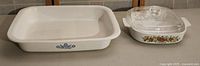 Two vintage CorningWare baking dishes displayed side by side on a shelf: rectangular white dish with blue cornflower pattern and oval white dish with L'Echalote pattern and glass lid.