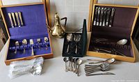 Overview of the lot showing three sets of flatware and serving utensils on table, two in wooden cases with fabric lining, one loose set, plus a decorative brass or metal pot.