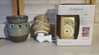 Fragrance warmers arranged side by side: green ceramic warmer, beige plug-in warmer with text, boxed ScentSationals warmer
