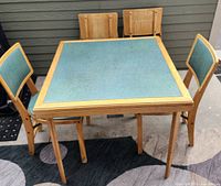 Square wooden card table with green padded vinyl top and four matching chairs with padded vinyl seats and backs clustered around it. The set is placed outdoors against a wall, showing overall condition and design.