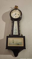 Full view of the banjo clock showing round clock face, decorated wooden body and framed nautical artwork on the base.